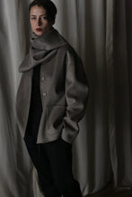 Load image into Gallery viewer, Set against a backdrop of draped fabric, a person wears a modern dark outfit paired with the Bamford Short Double-Face Cashmere in Undyed Natural, an oversized and warm gray scarf wrapped elegantly around their neck and shoulders. This luxurious cashmere piece enhances the ensemble&#39;s elegance, as soft lighting contributes to a moody, contemporary atmosphere.