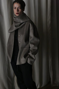 Set against a backdrop of draped fabric, a person wears a modern dark outfit paired with the Bamford Short Double-Face Cashmere in Undyed Natural, an oversized and warm gray scarf wrapped elegantly around their neck and shoulders. This luxurious cashmere piece enhances the ensemble's elegance, as soft lighting contributes to a moody, contemporary atmosphere.