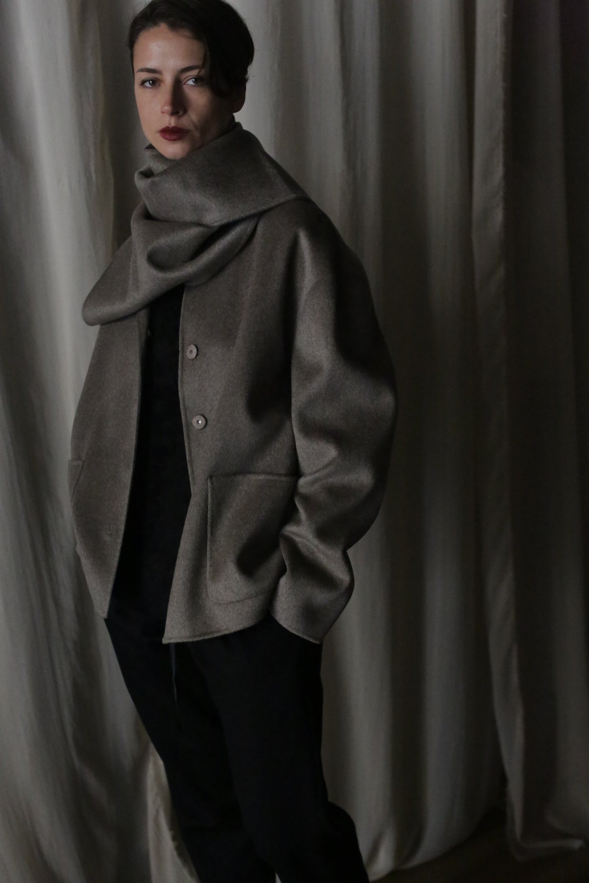 A person with short dark hair and bold lipstick wears a taupe oversized coat and an X Scarf - Double-face Cashmere | Undyed, standing before a softly draped fabric backdrop, gazing neutrally at the camera.