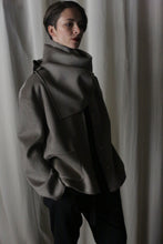 Load image into Gallery viewer, In a softly lit room, a person with short hair stands clad in the Bamford Short Double-Face Cashmere jacket in Undyed Natural. The modern, voluminous jacket with its distinctive large collar complements the serene white drapery backdrop as they gaze thoughtfully to the side, embodying a contemporary style.