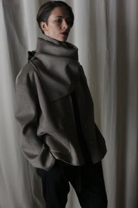 In a softly lit room, a person with short hair stands clad in the Bamford Short Double-Face Cashmere jacket in Undyed Natural. The modern, voluminous jacket with its distinctive large collar complements the serene white drapery backdrop as they gaze thoughtfully to the side, embodying a contemporary style.