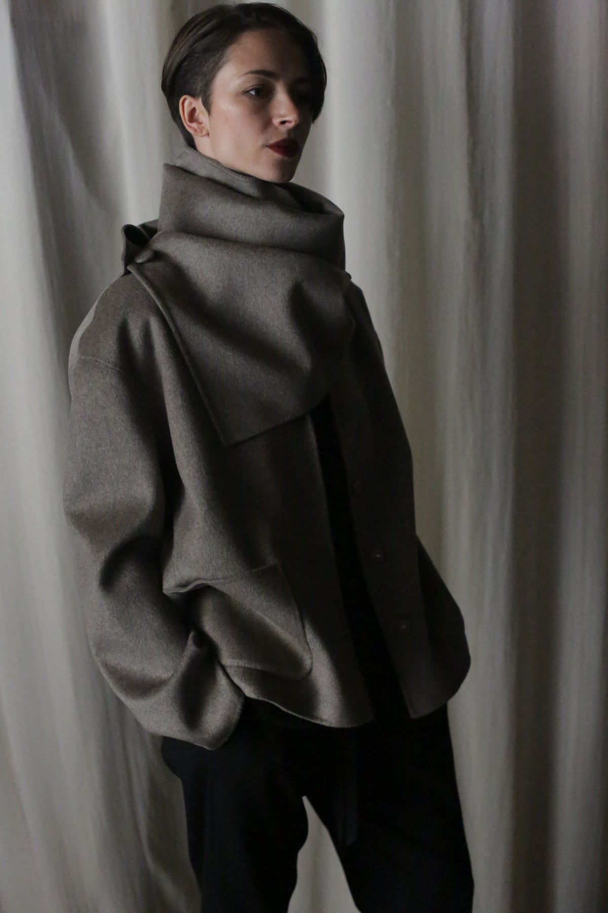 A person stands against a curtain, wearing the X Scarf crafted from versatile double-faced undyed cashmere. With short hair and hands in pockets, the dim lighting enhances the moody atmosphere.