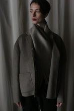 Load image into Gallery viewer, A person wrapped in a luxurious Bamford Short Double-Face Cashmere scarf in Undyed Natural and a dark gray coat stands against a neutral curtain backdrop. The dim lighting casts a moody ambiance, enhancing their contemplative expression as they gaze slightly off-camera, embodying modern elegance.