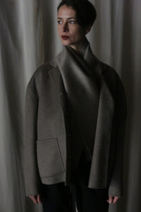 A person wrapped in a luxurious Bamford Short Double-Face Cashmere scarf in Undyed Natural and a dark gray coat stands against a neutral curtain backdrop. The dim lighting casts a moody ambiance, enhancing their contemplative expression as they gaze slightly off-camera, embodying modern elegance.