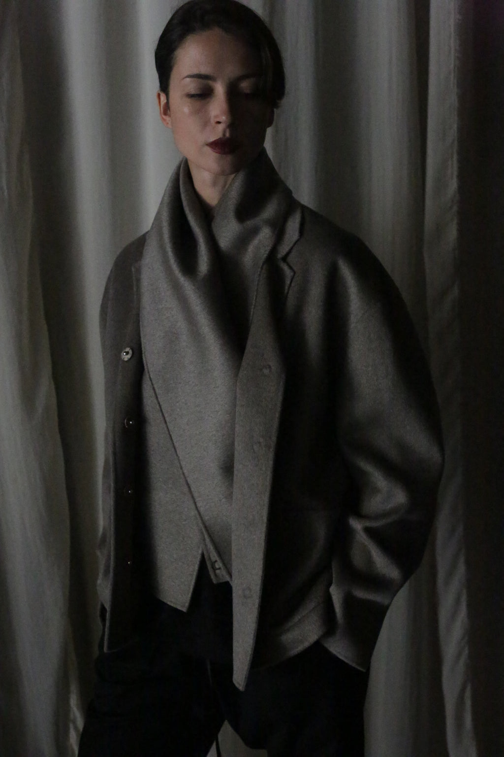 Eyes closed, a person stands draped in the X Scarf—Double-faced Cashmere and Undyed. This dark gray, oversized piece features a wide collar that complements their short hair and dark lipstick. Softly lit white curtains create a serene backdrop to this versatile layer.