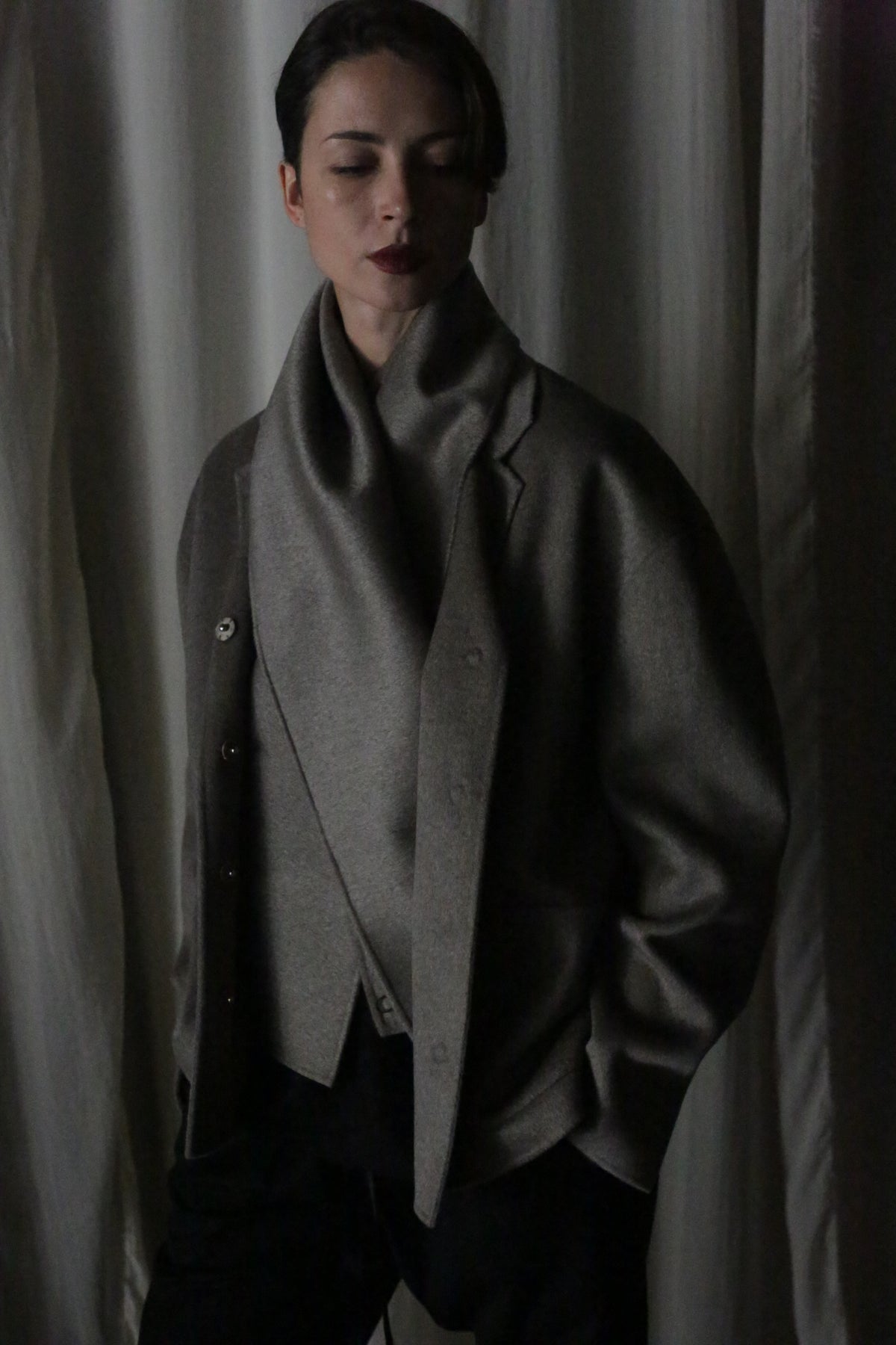 Eyes closed, a person stands draped in the X Scarf—Double-faced Cashmere and Undyed. This dark gray, oversized piece features a wide collar that complements their short hair and dark lipstick. Softly lit white curtains create a serene backdrop to this versatile layer.