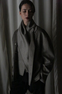 Wrapped in a Bamford Short Double-Face Cashmere scarf in Undyed Natural, a person stands in dim lighting, enveloped in a dark, oversized coat. The softly draped curtains create an elegant backdrop. With a calm expression, hands in pockets, and hair neatly parted, they embody a modern style.