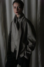 Load image into Gallery viewer, A person stands against a curtain backdrop, wearing the Bamford Short Double-Face Cashmere coat in Undyed Natural. This high-neck, oversized garment features an asymmetrical button design, crafted from luxurious cashmere that embodies modern style. Their expression is calm with one hand in their pocket while the dim lighting creates a moody atmosphere.