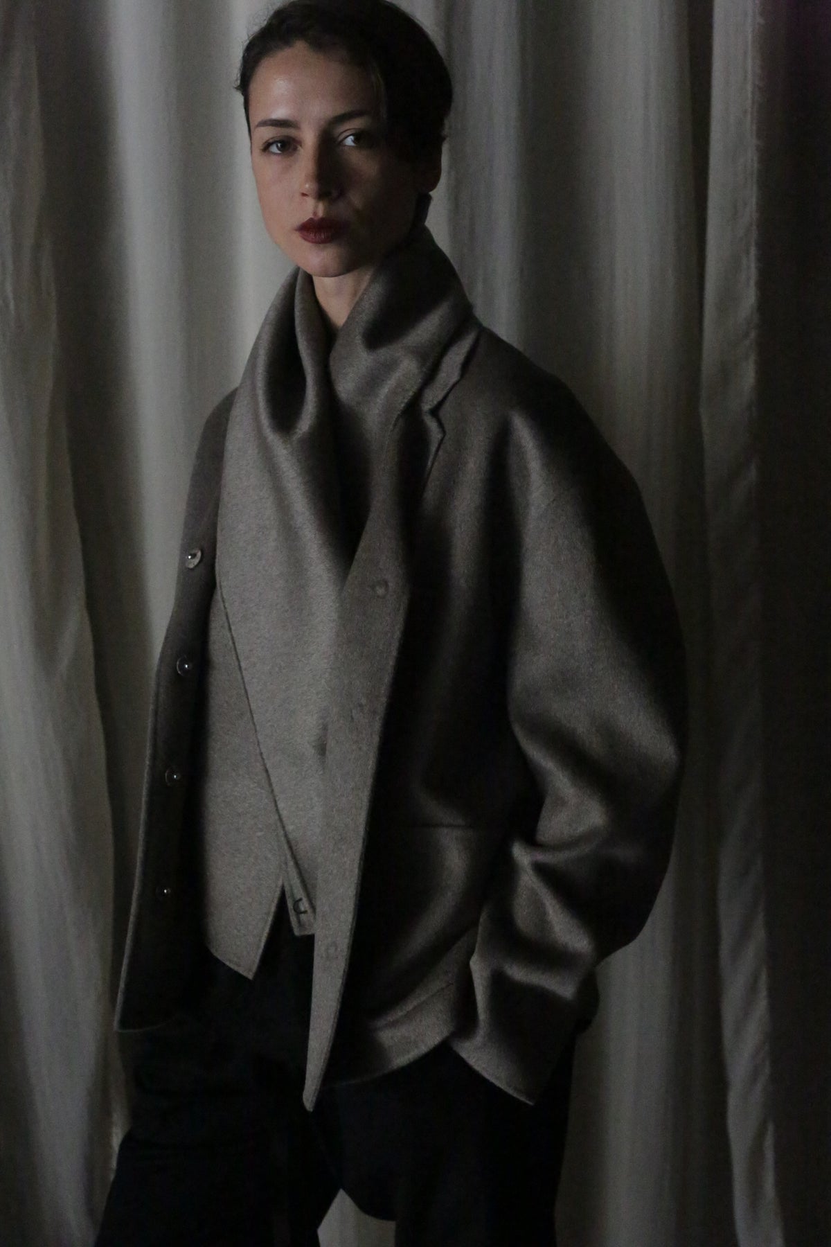 A person wears the X Scarf in double-faced, undyed cashmere, hands tucked in pockets against draped fabric. Dim lighting casts soft shadows, enhancing the moody atmosphere and highlighting the versatility of this chic accessory.