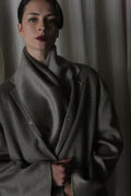 A person with short, dark hair and red lipstick stands against a neutral background, wrapped in an elegant X Scarf made of undyed double-face cashmere. The dim lighting accentuates the moody atmosphere, highlighting the elegance of sustainable fashion.