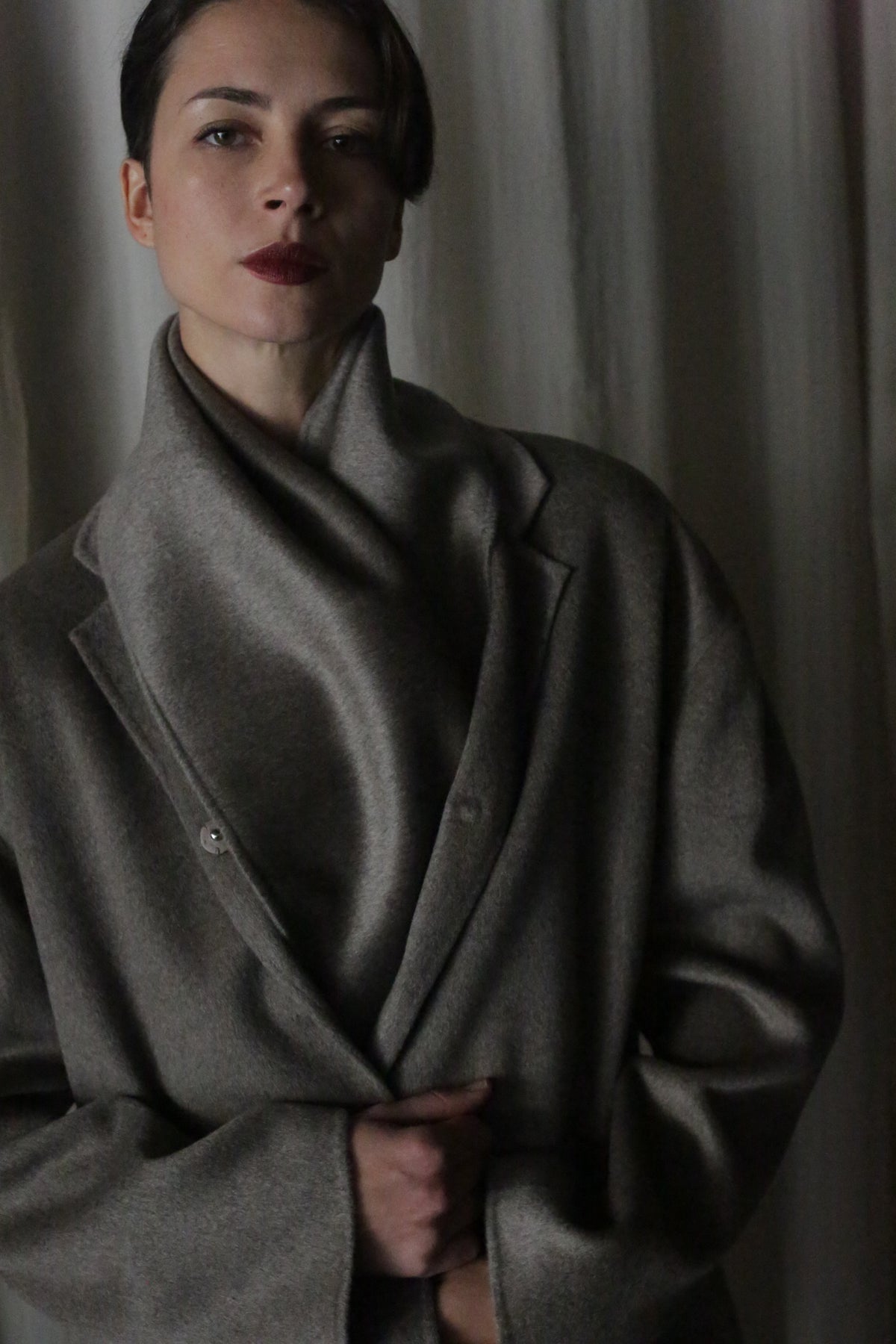A person with short, dark hair and red lipstick stands against a neutral background, wrapped in an elegant X Scarf made of undyed double-face cashmere. The dim lighting accentuates the moody atmosphere, highlighting the elegance of sustainable fashion.