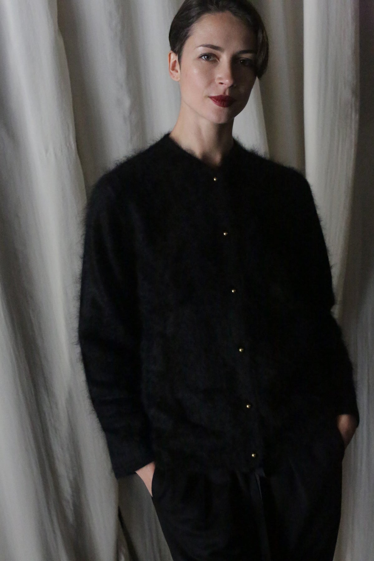 A person with short dark hair, dressed in the Brushed Cashmere Cardigan in Black and black pants, stands hands-in-pockets before white curtains, gazing at the camera with a slight smile—effortless style meets lightweight warmth.