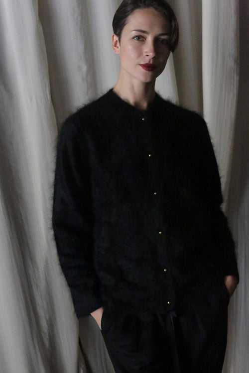 A person with short dark hair, dressed in the Brushed Cashmere Cardigan in Black and black pants, stands hands-in-pockets before white curtains, gazing at the camera with a slight smile—effortless style meets lightweight warmth.