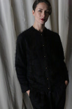 Load image into Gallery viewer, A person with short hair and red lipstick wears a luxurious black Brushed Cashmere Cardigan and dark pants, standing against a light draped background. Their hands are in their pockets, looking at the camera.