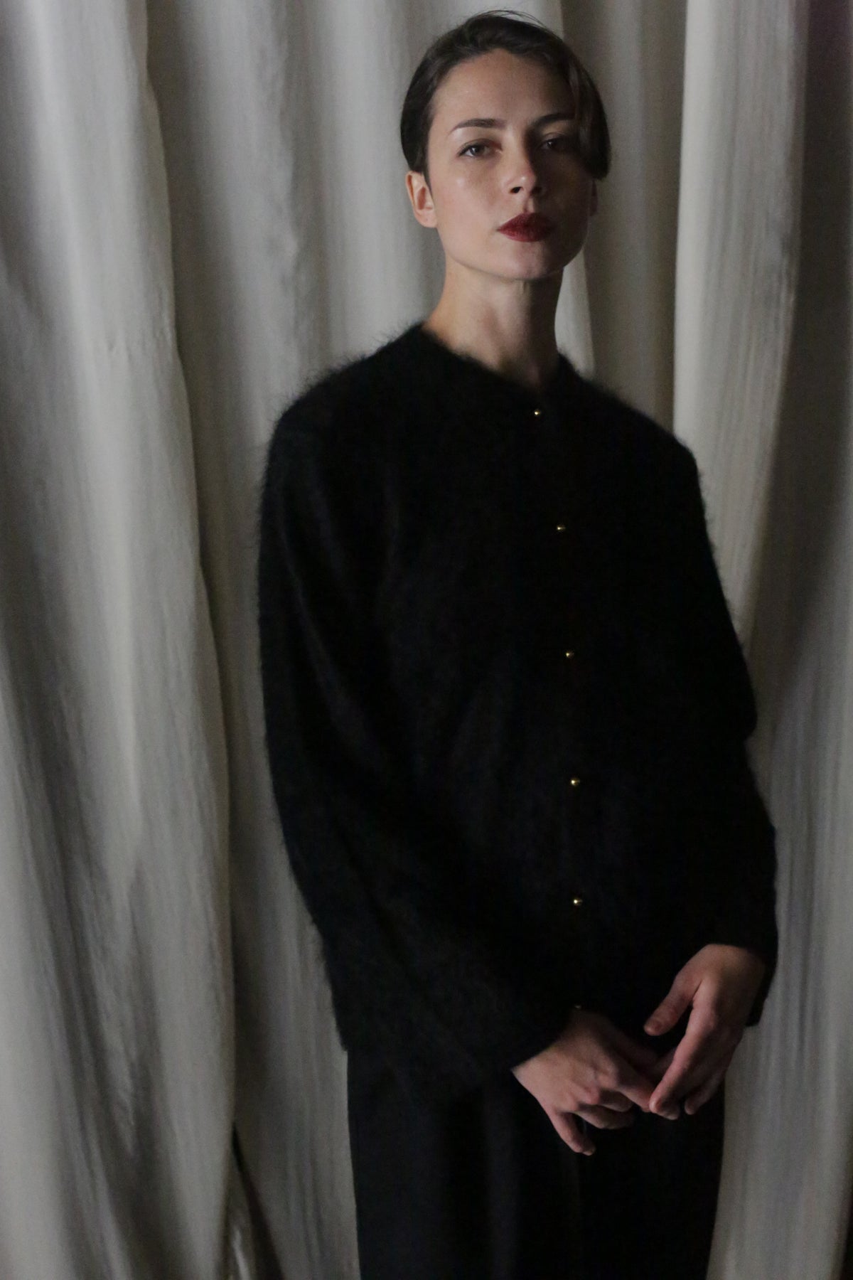 A woman with short dark hair stands against cream curtains, wearing the Brushed Cashmere Cardigan in Black with gold buttons. She gazes at the camera with a neutral expression, hands clasped, showcasing the cardigan’s soft, lightweight warmth.