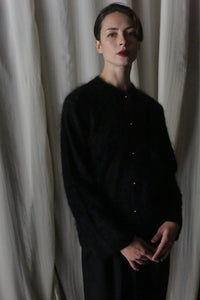 A short-haired person stands before a draped curtain, hands gently clasped, wearing a Brushed Cashmere Cardigan and pants. The dim lighting casts soft shadows, highlighting the elegance of their luxury knitwear ensemble.