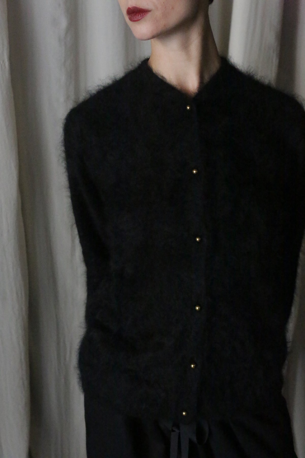 Someone stands in front of a light curtain, wearing the Brushed Cashmere Cardigan in Black with matching black pants. Their head is slightly turned, revealing only part of their face.