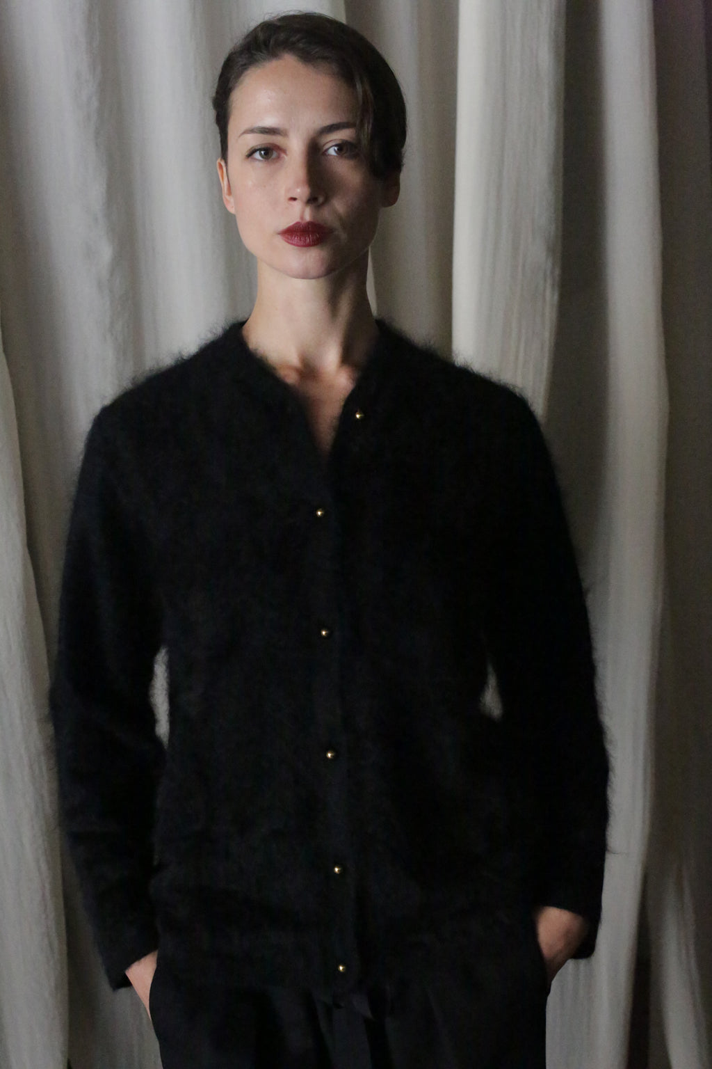 A woman with short brown hair stands against cream curtains, wearing the Brushed Cashmere Cardigan in black and matching pants. Hands in her pockets, she gazes at the camera with a neutral expression, embodying its lightweight warmth.
