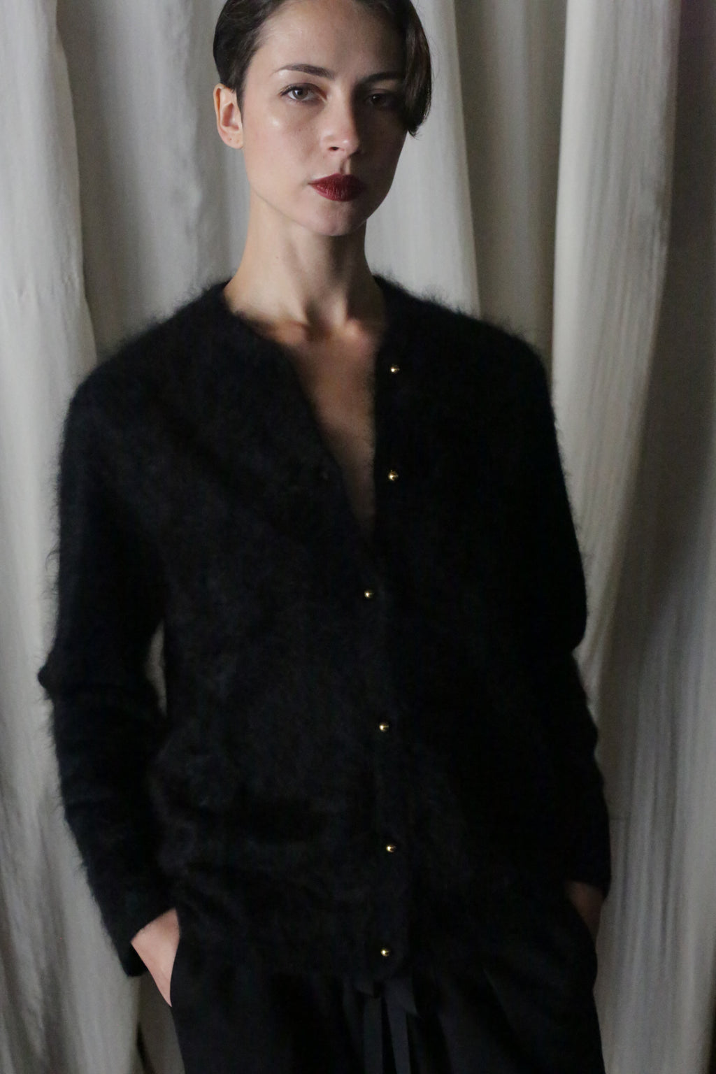 A person with short, dark hair stands before pale drapes, hands in pockets and a neutral expression, effortlessly showcasing the lightweight warmth of the Brushed Cashmere Cardigan | Black paired with dark pants.