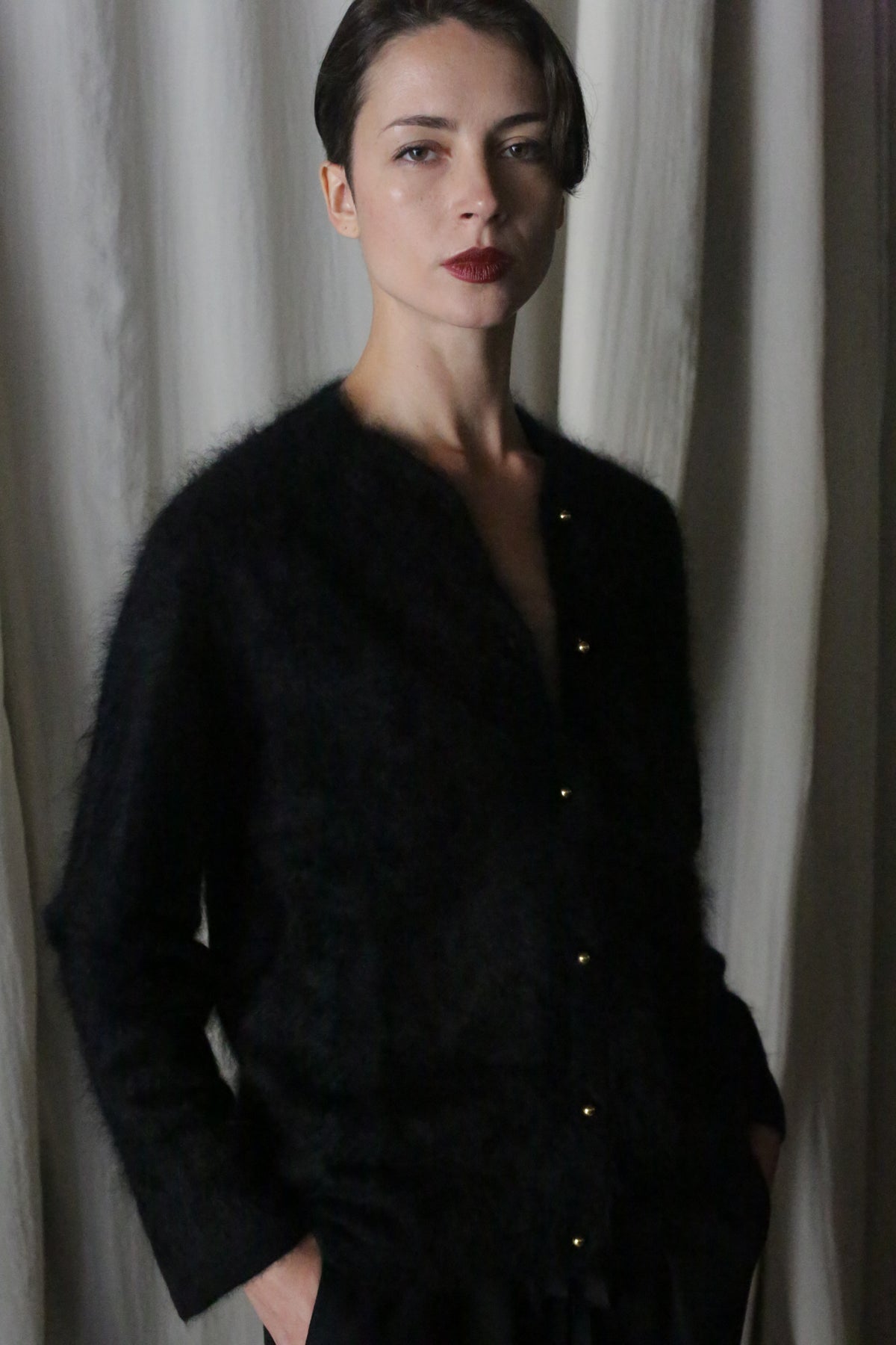 A person with short dark hair stands before light curtains, hands in pockets, confidently modeling the Brushed Cashmere Cardigan | Black—a stylish piece that exudes lightweight warmth.