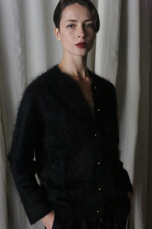 A person with short dark hair stands before light curtains, hands in pockets, confidently modeling the Brushed Cashmere Cardigan | Black—a stylish piece that exudes lightweight warmth.