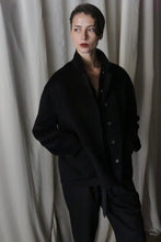 Load image into Gallery viewer, A person with short dark hair stands against a backdrop of white curtains, clad in the Bamford Short Double-Face Cashmere in black. They have a contemplative expression, hands tucked in their pockets. The soft lighting lends a dramatic effect, making this contemporary wardrobe essential truly captivating.