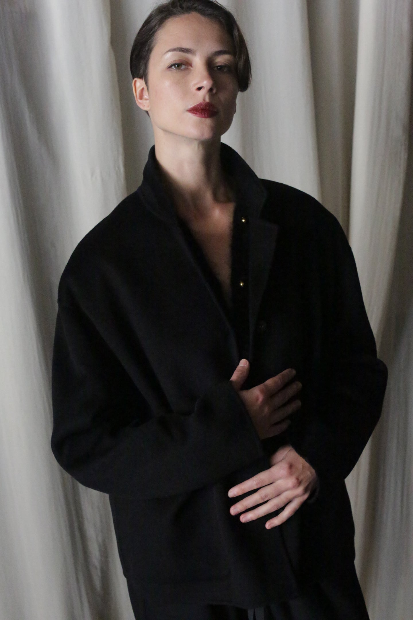 A person elegantly poses in front of light-colored curtains, wearing the Bamford Short Double-Face Cashmere coat in black. One hand gently holds the coat closed while the other rests near the waist, capturing the essence of a contemporary wardrobe essential.