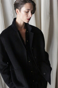 Clad in a must-have modern wardrobe piece—the Bamford Short Double-Face Cashmere in Black—a person stands against a delicately textured backdrop. With their head gently inclined downward and hands nestled into the opulent double-faced cashmere pockets, they embody a contemplative yet relaxed stance.