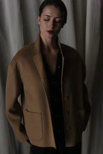 Load image into Gallery viewer, Dressed in a Bamford Short Double-Face Cashmere coat in camel, a woman stands with her eyes closed, enveloped in luxury over a chic black ensemble. The softly lit backdrop and white curtains augment the tranquil elegance, embodying a modern wardrobe essential.