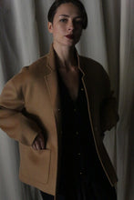 Load image into Gallery viewer, A person with short dark hair stands confidently, wearing a Bamford Short Double-Face Cashmere coat in Camel over a black outfit. This contemporary wardrobe essential is expertly crafted to provide elegance, complemented by the soft, subdued atmosphere created by the light-colored curtains in the background.