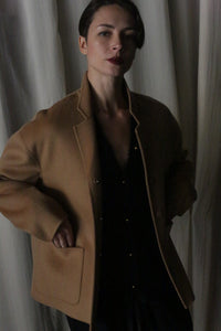 A person with short dark hair stands confidently, wearing a Bamford Short Double-Face Cashmere coat in Camel over a black outfit. This contemporary wardrobe essential is expertly crafted to provide elegance, complemented by the soft, subdued atmosphere created by the light-colored curtains in the background.