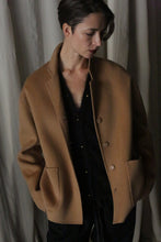 Load image into Gallery viewer, A person wearing a Bamford Short Double-Face Cashmere coat in Camel over a black outfit stands with eyes looking downward, hands in pockets, in front of a cream-colored curtain. The double-faced cashmere and soft lighting cast gentle shadows, creating an intimate and contemplative mood.