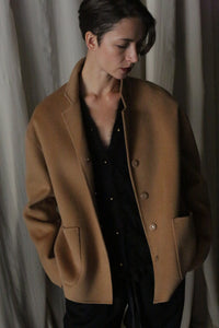 A person wearing a Bamford Short Double-Face Cashmere coat in Camel over a black outfit stands with eyes looking downward, hands in pockets, in front of a cream-colored curtain. The double-faced cashmere and soft lighting cast gentle shadows, creating an intimate and contemplative mood.