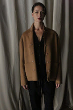 Load image into Gallery viewer, A person with dark hair stands against a draped, neutral-colored background, wearing a camel Bamford Short Double-Face Cashmere coat over a black outfit. This coat is a contemporary wardrobe essential. Their expression is serious and composed.