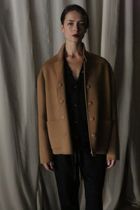 A person with dark hair stands against a draped, neutral-colored background, wearing a camel Bamford Short Double-Face Cashmere coat over a black outfit. This coat is a contemporary wardrobe essential. Their expression is serious and composed.