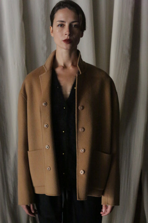 In front of a curtain, a person stands wearing the Bamford Short Double-Face Cashmere coat in camel, layered over a black outfit. Their short hair and neutral expression complement the contemporary elegance of this luxurious piece, while the soft and subdued lighting enhances its appeal.