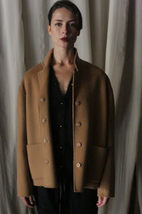 In front of a curtain, a person stands wearing the Bamford Short Double-Face Cashmere coat in camel, layered over a black outfit. Their short hair and neutral expression complement the contemporary elegance of this luxurious piece, while the soft and subdued lighting enhances its appeal.