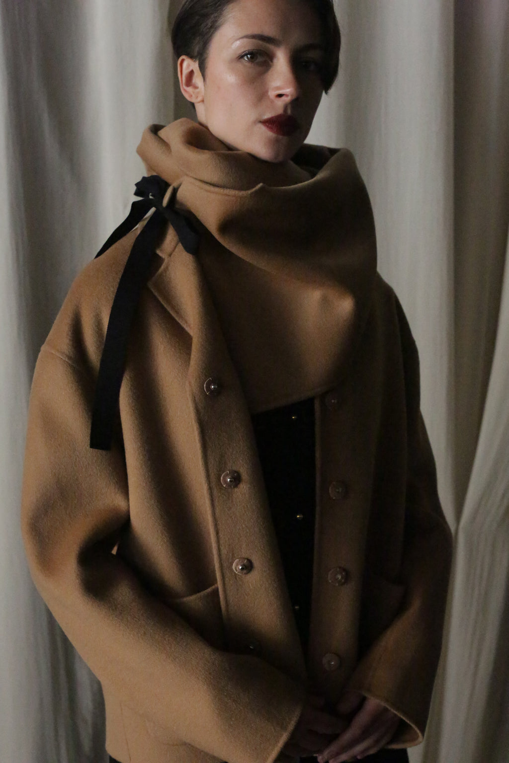 A person wraps a large X Scarf - Double-face Cashmere in Camel partly over their mouth, featuring a black ribbon on the shoulder, against a softly lit curtain.