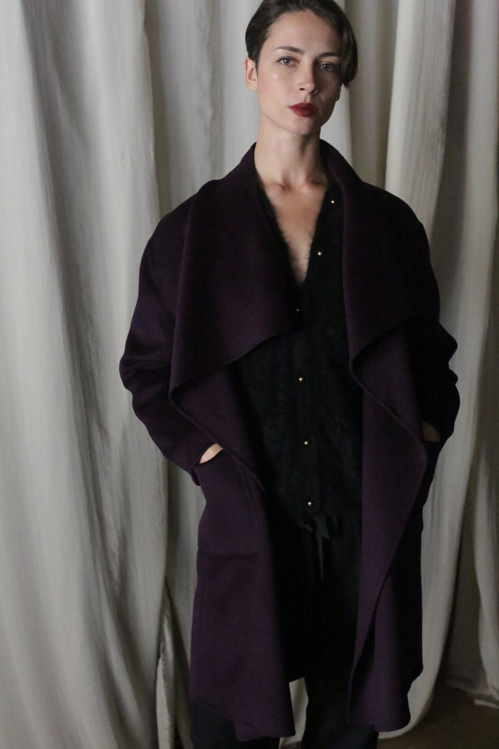 A person with short hair stands in front of a curtain, wearing the Winston Long Double-face coat in aubergine over a black outfit. Their hands are in their pockets, and they gaze at the camera with a neutral expression, embodying versatile styling options.