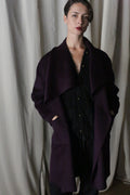 A person with short hair stands in front of a curtain, wearing the Winston Long Double-face coat in aubergine over a black outfit. Their hands are in their pockets, and they gaze at the camera with a neutral expression, embodying versatile styling options.