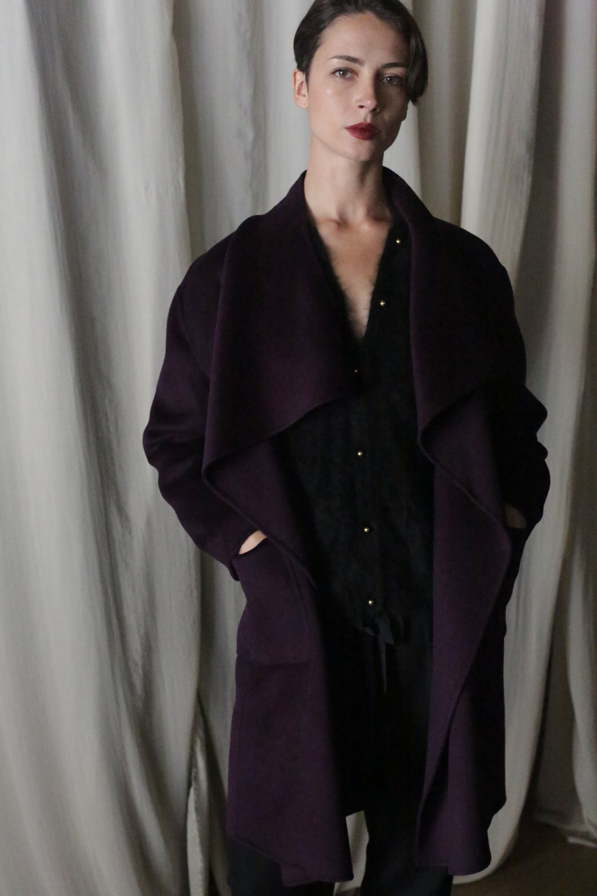 A person with short hair stands in front of a curtain, wearing the Winston Long Double-face coat in aubergine over a black outfit. Their hands are in their pockets, and they gaze at the camera with a neutral expression, embodying versatile styling options.