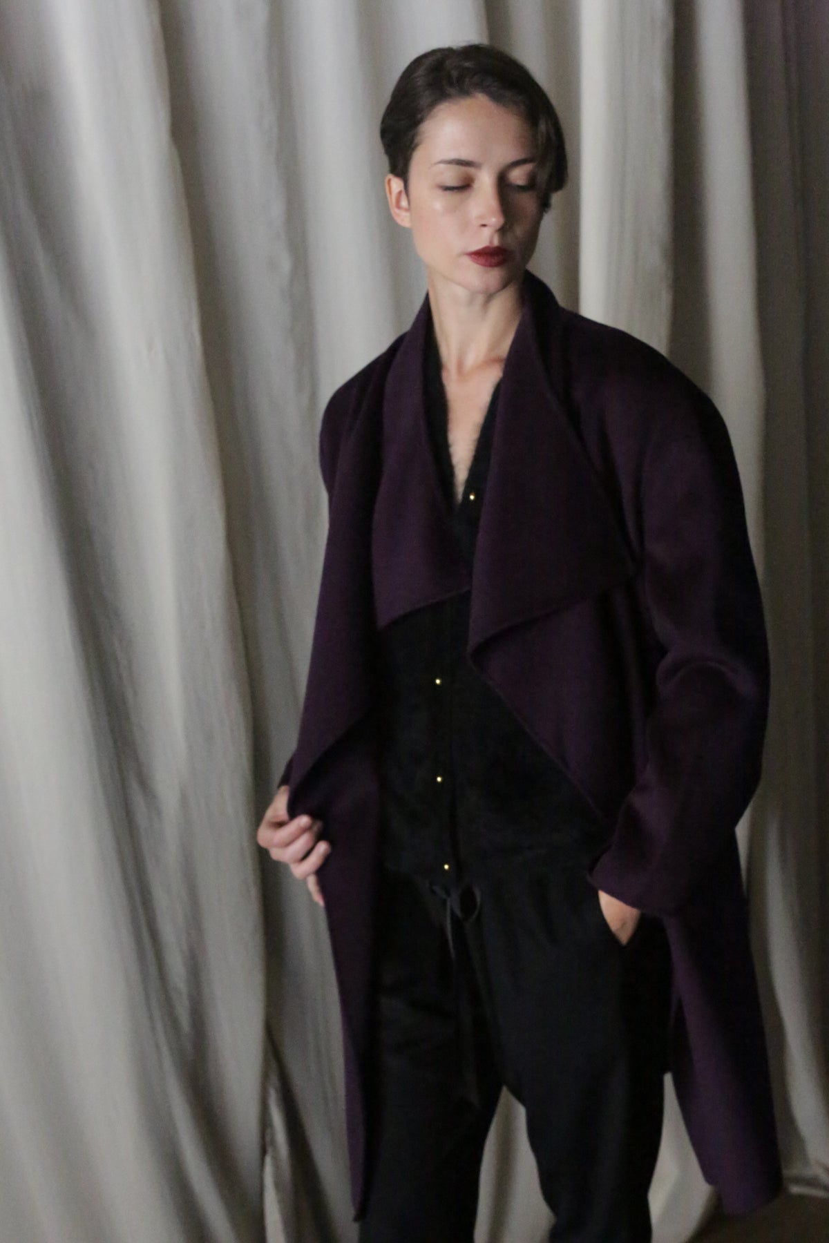 A person with short dark hair stands in front of draped fabric, wearing an aubergine Winston Long Double-face coat over a black outfit. The sophisticated double-faced cashmere accentuates their contemplative expression as they look slightly downward.