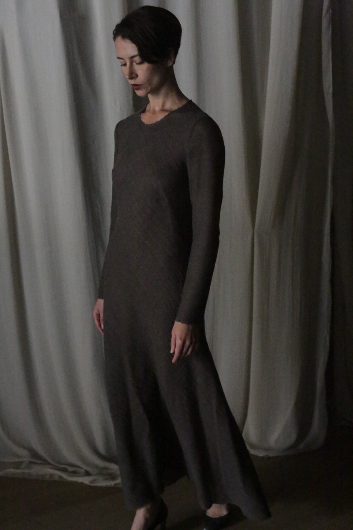 A woman with short dark hair stands indoors, wearing the Long Sleeved Crewneck Dress in dark brown wool gauze. She gazes downward calmly, framed by softly draped beige curtains—handmade in San Francisco.