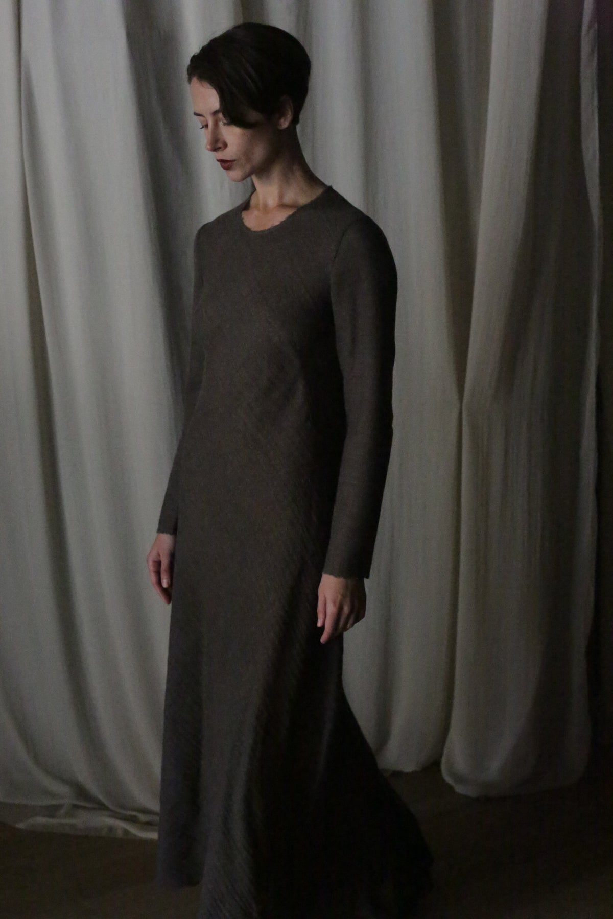 A woman with short hair stands in a dim room, wearing the Long Sleeved Crewneck Dress. Flowing drapes hang behind her as she gazes downward with a calm expression, highlighting the dress’s timeless handmade San Francisco elegance.