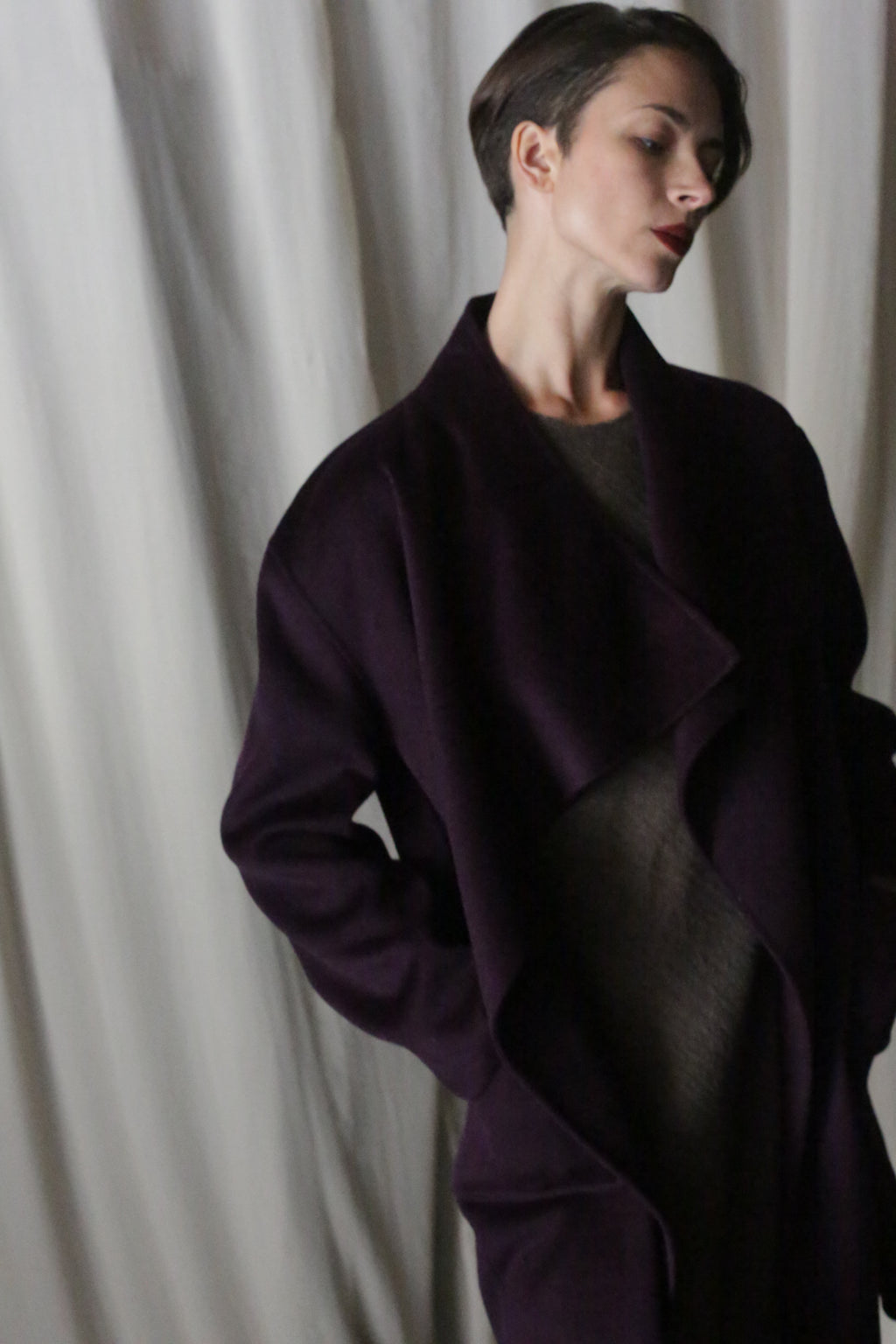 A person with short hair poses elegantly against a white curtain in an aubergine Winston Long Double-face coat. The luxurious double-faced cashmere garment offers versatile styling options as they gaze thoughtfully to the side.