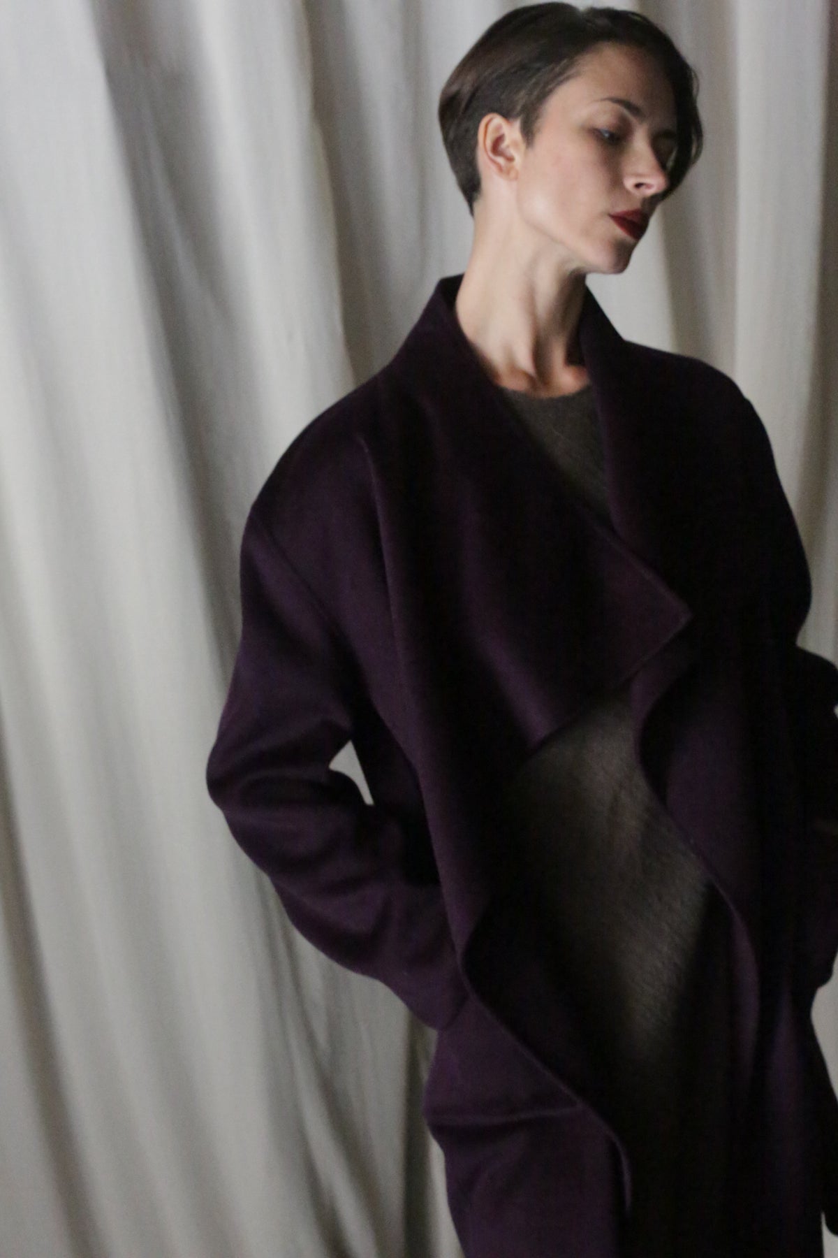 A person with short hair poses elegantly against a white curtain in an aubergine Winston Long Double-face coat. The luxurious double-faced cashmere garment offers versatile styling options as they gaze thoughtfully to the side.