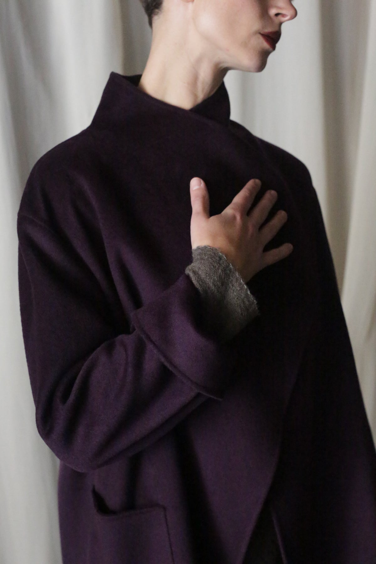 Against a light curtain, a person wears the Winston Long Double-face coat in aubergine with a high collar made of double-faced cashmere. Their hand rests on their chest, and they have short hair. Soft lighting enhances the calm atmosphere, highlighting the coat's versatile styling options.
