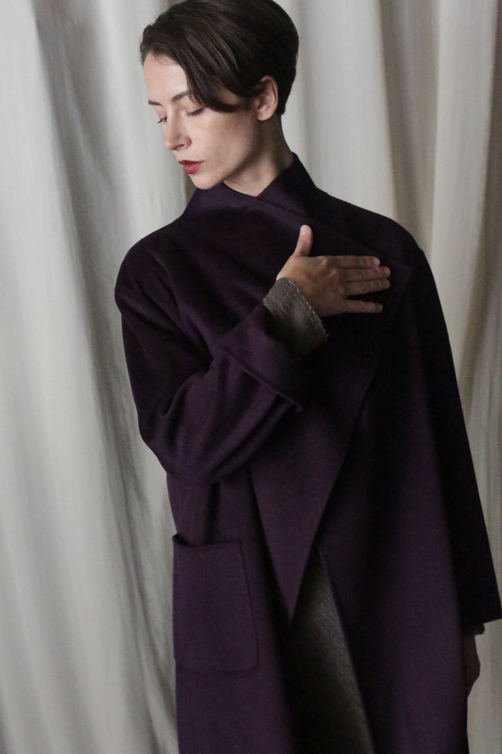 Against a light curtain backdrop, a person with short hair wears an aubergine Winston Long Double-face coat over a gray sweater. With their right hand on their chest and gazing thoughtfully downwards, they showcase versatile styling options in every detail.