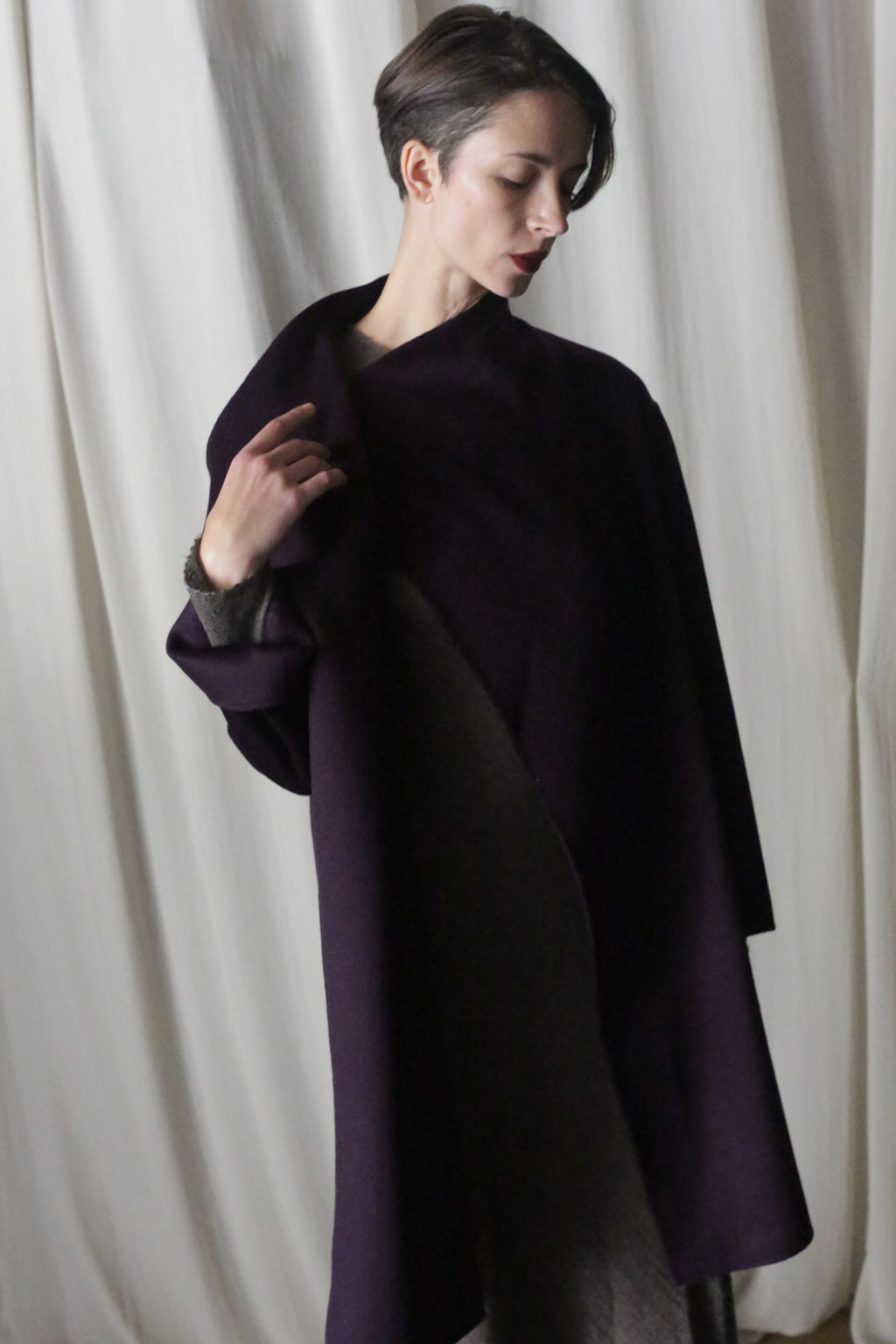 With short dark hair, a person in a Winston Long Double-face coat in Aubergine stands against a soft background. The double-faced cashmere piece offers versatile styling, while they gaze downward with serenity.