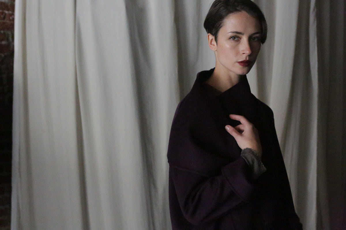 A person with short hair stands before draped curtains, wearing the Winston Long Double-face coat in Aubergine. They hold part of the coat contemplatively, while soft lighting highlights its versatility.